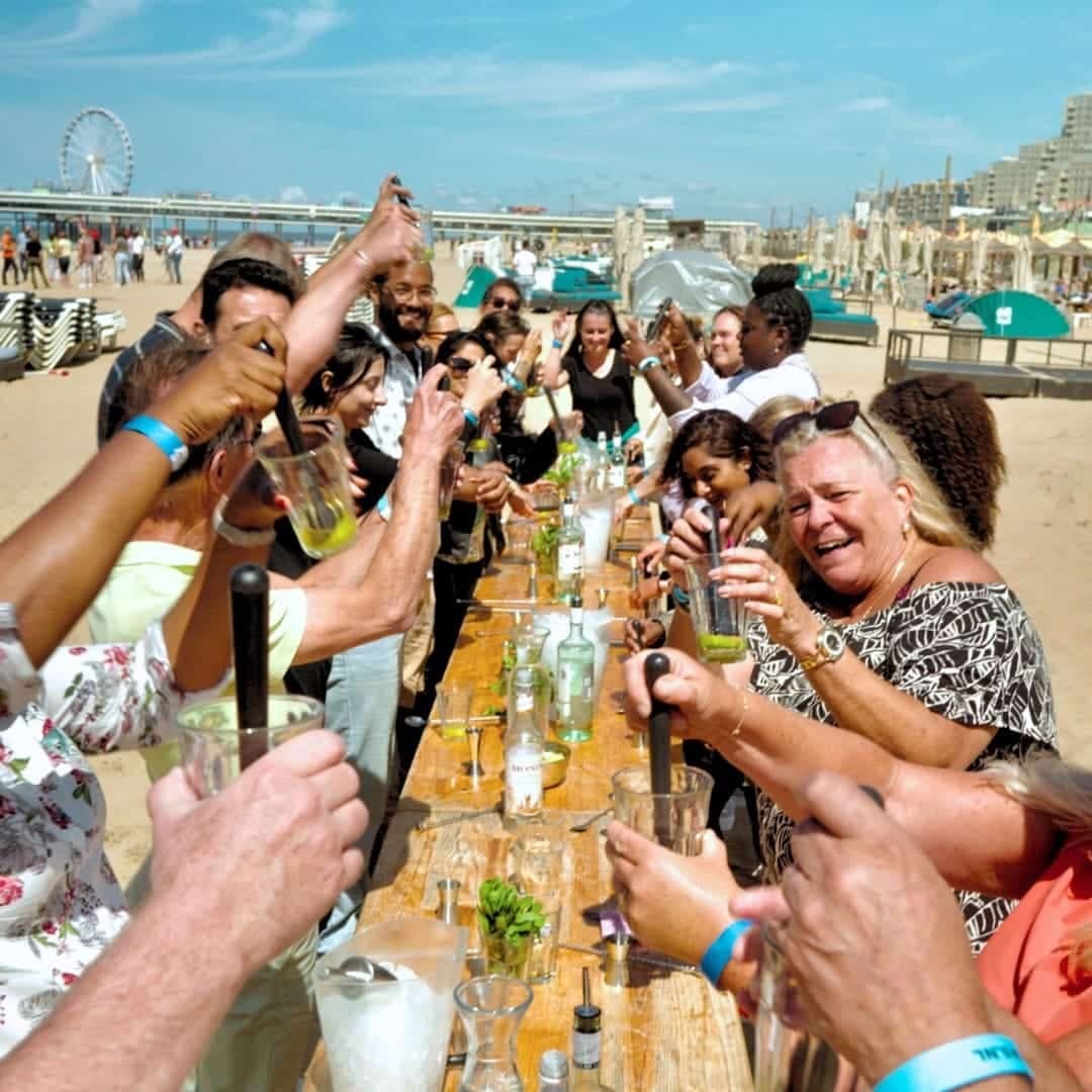 Groep shaken cocktails aan een lange tafel op het strand van Scheveningen, met pier en reuzenrad